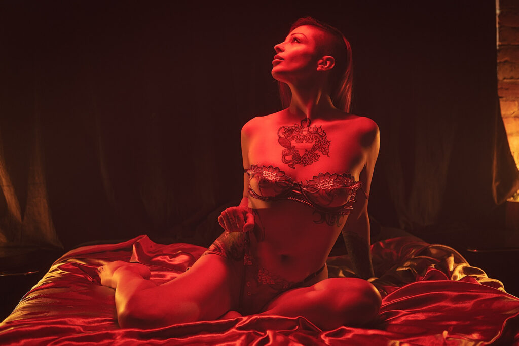 A woman wearing lingerie sits on a bed under red lighting, with tattoos visible on her chest and arm, looking upwards—I love to tease and be teased.