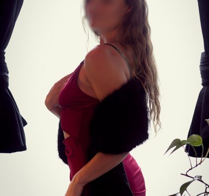 A goddess personified, a figure in a red dress and black shawl stands by a window with dark curtains and a plant, facing sideways. Face is blurred.