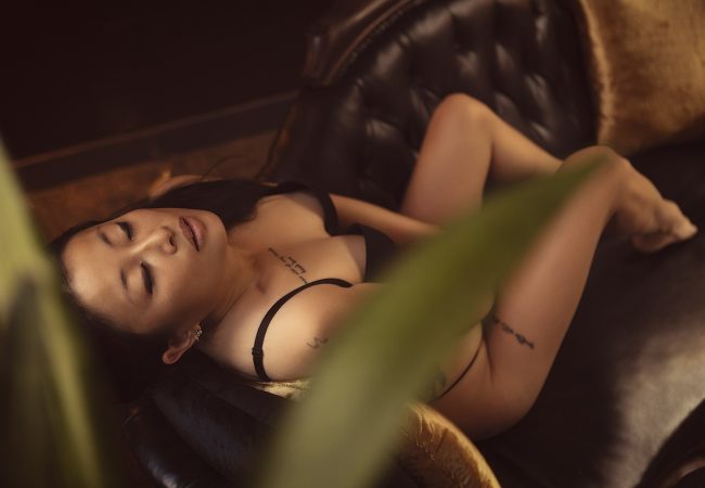 A woman in black lingerie, your ideal Korean companion, reclines on a dark tufted couch with her eyes closed; green leaves are visible in the foreground.