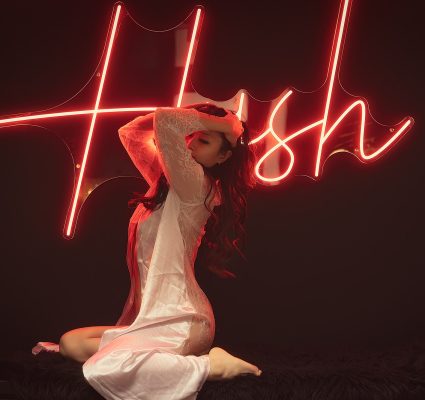 A woman in a sheer white dress, your ideal Korean companion, poses on her knees in front of a neon sign that reads "Hush" in red script.