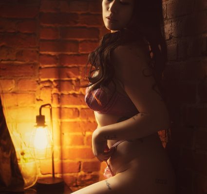 A woman in pink lingerie stands against a brick wall, illuminated by a nearby vintage lamp—your ideal Korean companion in an intimate, dreamy setting.