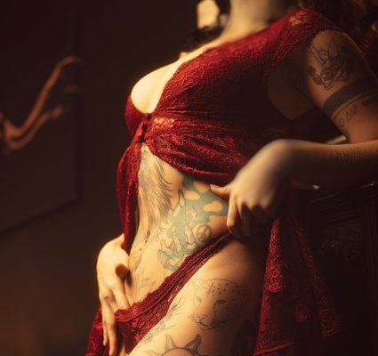 A person in a red lace outfit poses in a dimly lit room, displaying multiple tattoos and piercings on their body. Their face is blurred, hinting at their unique style—I have many tattoos and even a split tongue.
